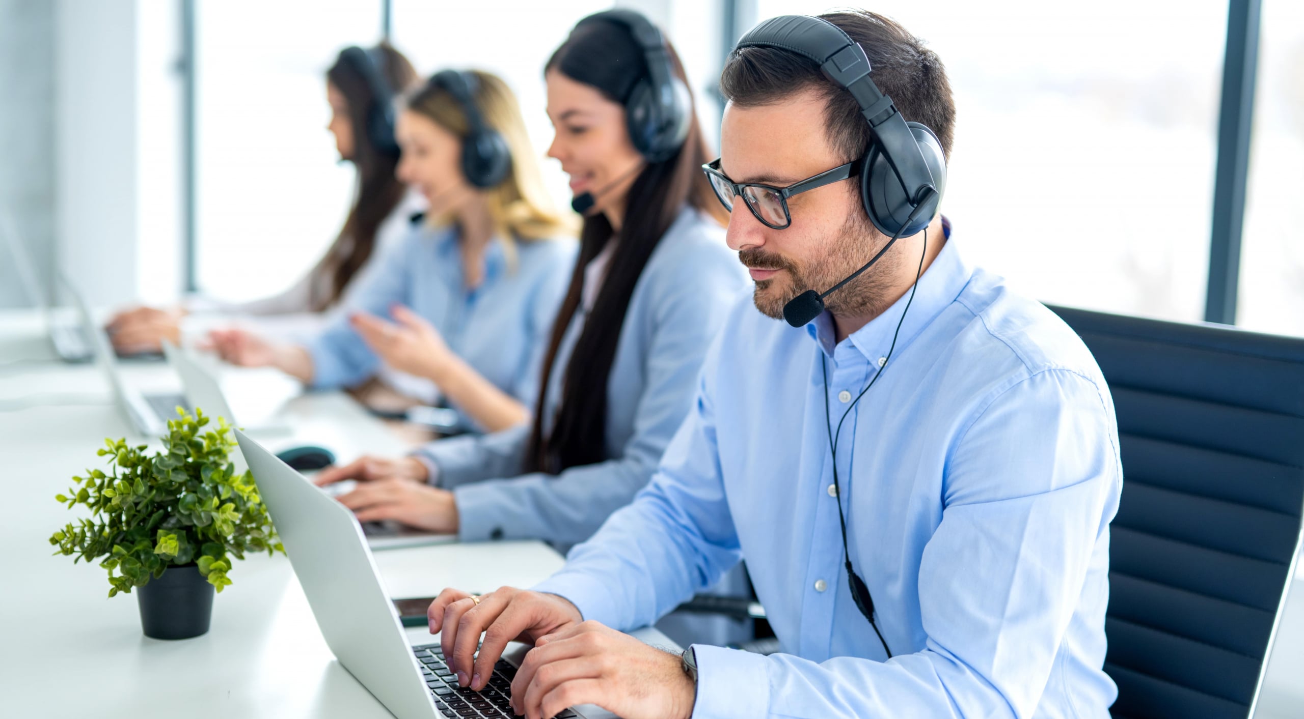 Image2_callcenter_worker_sitting _in_row-min Call center agents wearing headsets and typing on laptops at a desk.