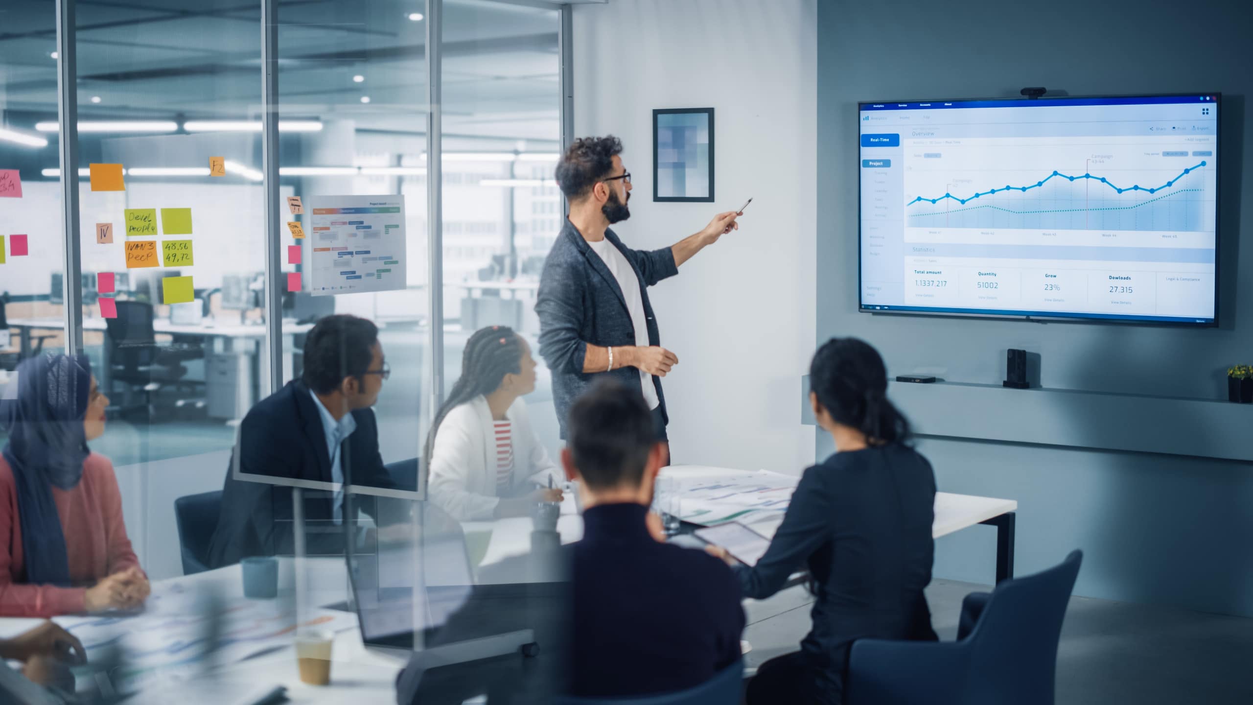Image3_man_pointing_at_graph_contactcenter_meeting-min A group of professionals sitting at a desk in a meeting led by a male pointing at a screen with a chart.