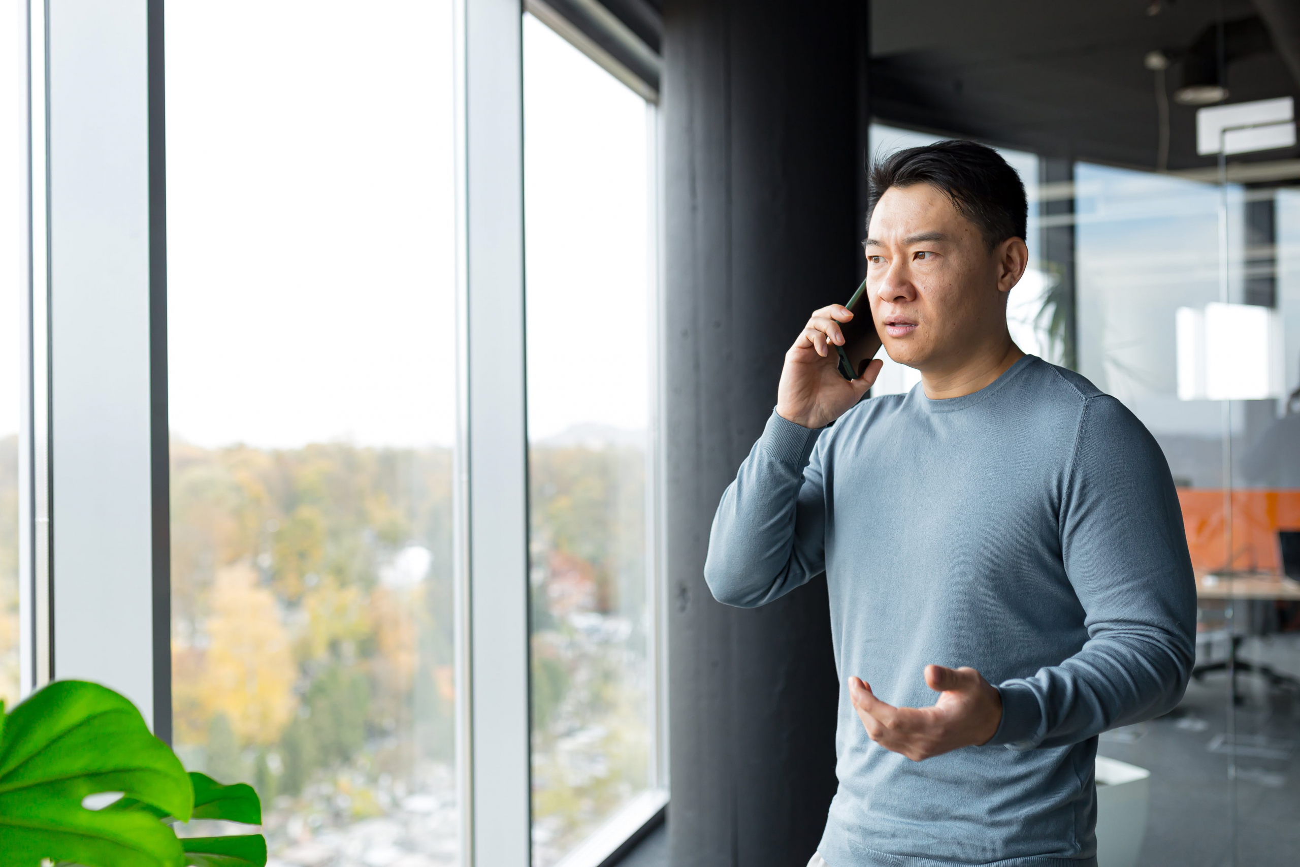 Asian Man looking out the window of his office on his cell phone concerned