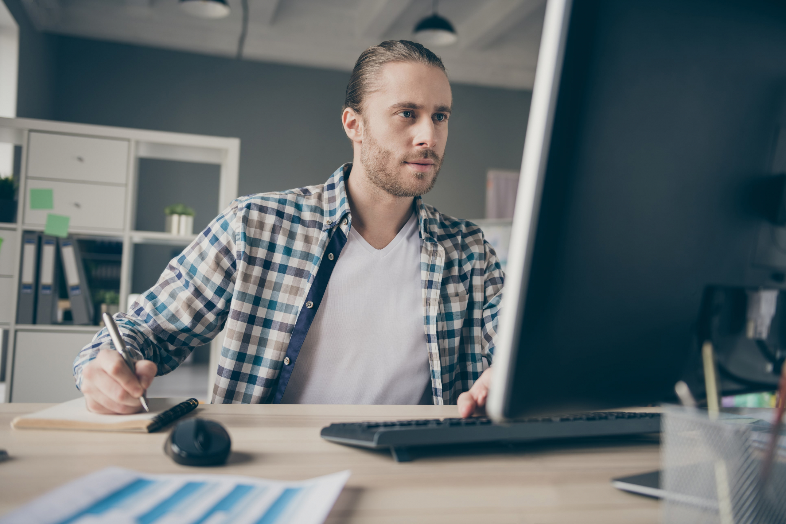 Bearded male salesperson writing down notes sitting at his computer