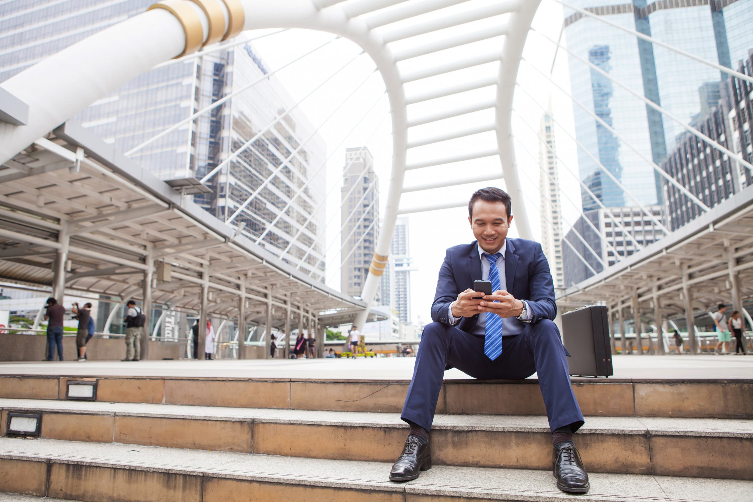 Young business man sitting outside on steps smiling and texting on his cell phone
